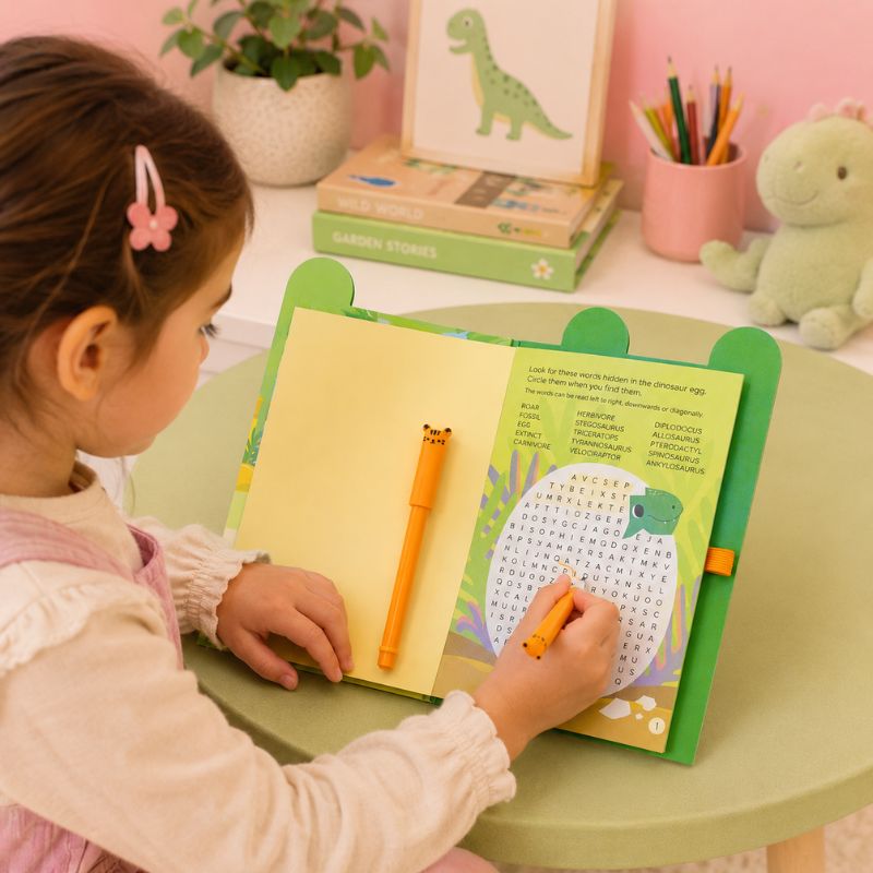 Child using a green children's desk with a Dinosaur Busy Whiz book and writing tools on a pink wall background - Loveshack Giftware