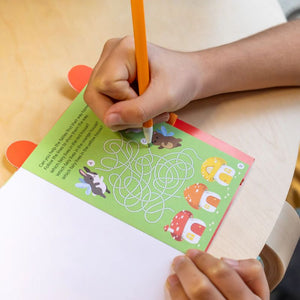 Hand holding a pencil over a coloring book with a maze and cartoon animals on a white surface.