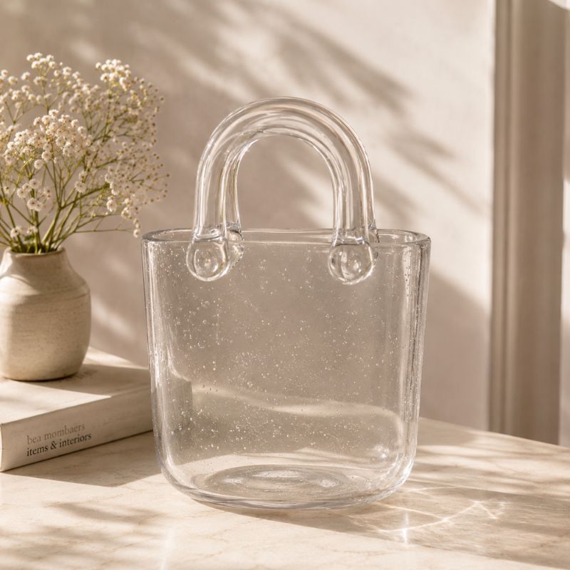 Clear glass tote bag on a light surface with a vase of flowers and books in the background.