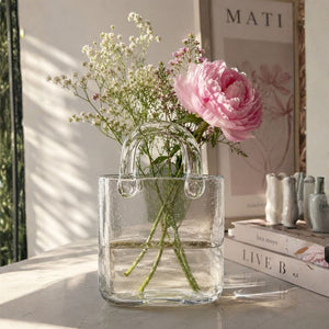 Clear glass vase shaped like a handbag with flowers inside, placed on a surface.