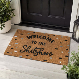 A rectangular coir doormat with the text 'WELCOME TO THE Shitshow' in black lettering, decorated with small black, gray, and white speckles, shown on a light gray wooden floor in front of a black door with potted plants and a lantern nearby.-Welcome to the Shitshow Doormat Black