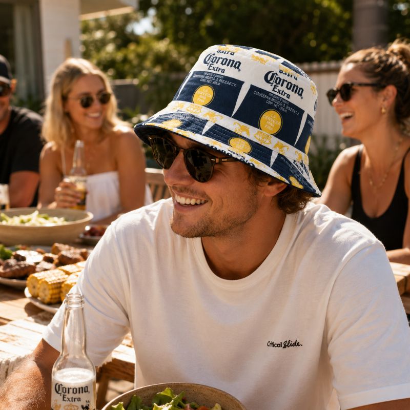 A man wearing a white t-shirt and a Corona Party Bucket Hat Reversable
 with a navy blue and yellow Corona Extra pattern, sitting at a table with friends and a beer bottle in a sunny outdoor setting.