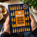 Book titled '101 Whiskies to Try Before You Die' held by a person with a glass of whiskey and snacks in the background.