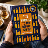 Book titled '101 Whiskies to Try Before You Die' held by a person with a glass of whiskey and snacks in the background.