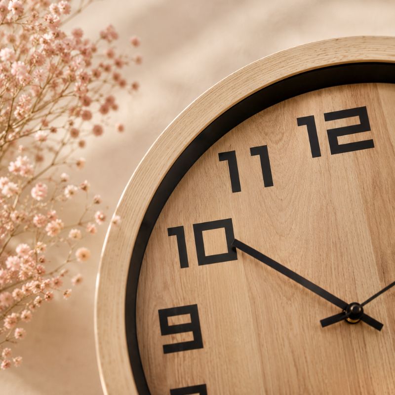 Close-up of a 40cm timber-look wall clock with a wood face and frame, black numerals, and black hands, styled with soft pink dried flowers against a warm beige background.