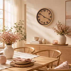 A 40 cm Timber Look Wall Clock with a light wood frame and black numerals hangs on a beige wall in a softly lit dining room. The scene includes a wooden table, chairs, vases with pink flowers, books, and candles, creating a warm, inviting interior.
