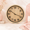 A 40cm Timber Look Wall Clock with a light natural wood grain face and frame, black numerals, and black hands, styled on a light surface with pink decorative props in the background.