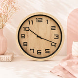 A 40cm Timber Look Wall Clock with a light natural wood grain face and frame, black numerals, and black hands, styled on a light surface with pink decorative props in the background.