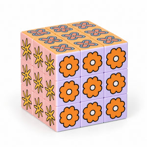 A 3x3x3 Art Cube – Flower Pop puzzle cube with a retro floral pattern, featuring orange flowers on the front, yellow flowers on the left, and a purple top with orange and blue geometric designs, set against a plain white background.