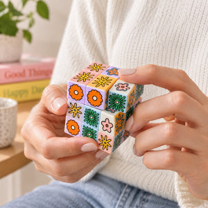 A person in a white knit sweater holds a Art Cube – Flower Pop small square puzzle cube decorated with colorful floral patterns in orange, green, yellow, and pink. The background shows a cozy indoor setting with a plant, books, and a mug.