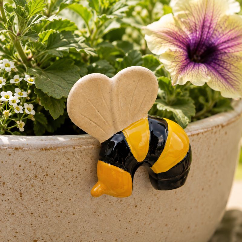 A ceramic bee garden stake or planter topper with a beige heart-shaped top and black and yellow body, shown leaning against green plants in a speckled beige pot.-Bee Pot Hanger Black Yellow -  7cm