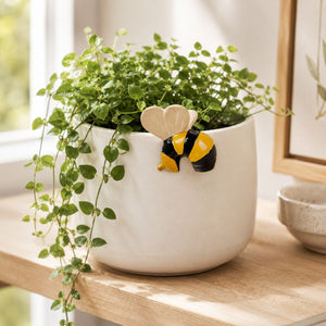 A white ceramic planter filled with trailing green ivy, decorated with a small yellow and black bee figure and a beige heart-shaped topper, displayed on a wooden shelf in a bright indoor setting.-Bee Pot Hanger Black Yellow -  7cm