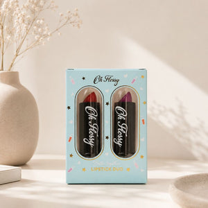 A boxed lipstick duo set labeled 'Oh Flossy' standing on a tabletop. The light blue package contains two lipsticks visible through oval windows, one red and one purple.