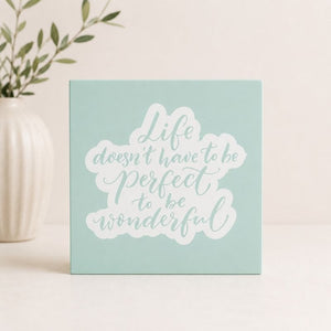 Motivational card with 'Life doesn't have to be perfect to be wonderful' text on a light blue background, placed next to a vase with greenery.