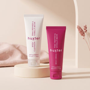 Huxter Body Cleanser and Body Cream tubes displayed on warm ivory pedestal