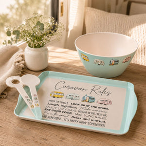 A styled tabletop scene showing a light aqua rectangular serving tray with a printed 'Caravan Rules' sign, accompanied by two matching white spoons and a matching light aqua bowl. The setup is on a wooden table with a vase of baby's breath and a cozy neutral background.