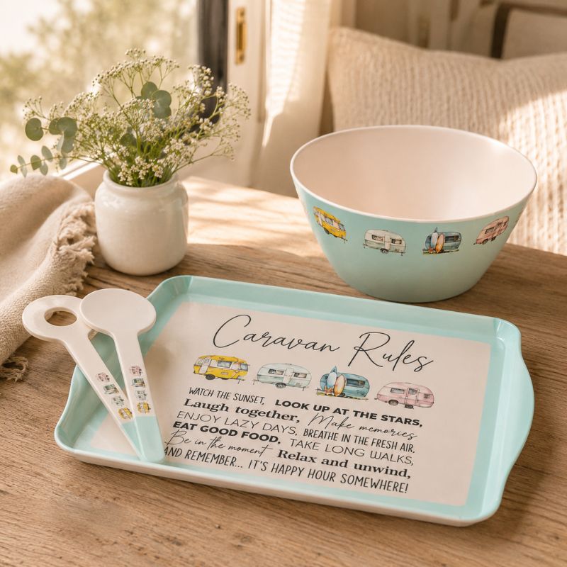 A styled tabletop scene showing a light aqua rectangular serving tray with a printed 'Caravan Rules' sign, accompanied by two matching white spoons and a matching light aqua bowl. The setup is on a wooden table with a vase of baby's breath and a cozy neutral background.
