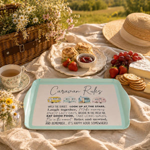 A rectangular teal tray with a white printed sign reading 'Caravan Rules' is styled on a picnic blanket outdoors. A straw hat, woven basket, flowers, tea cup, strawberries, grapes, biscuits, and cheese surround it, creating a real outdoor picnic environment.