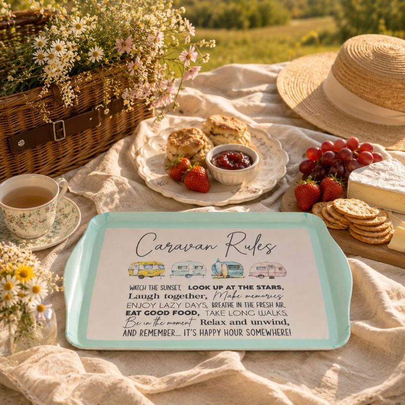 A rectangular teal tray with a white printed sign reading 'Caravan Rules' is styled on a picnic blanket outdoors. A straw hat, woven basket, flowers, tea cup, strawberries, grapes, biscuits, and cheese surround it, creating a real outdoor picnic environment.