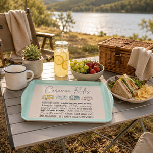 A Caravan Bamboo Melamine Serving Tray with a mint green border and a white printed 'Caravan Rules' picnic sign is placed on an outdoor folding table beside a sandwich, grapes, a drink, a wicker picnic basket, and a lake setting.