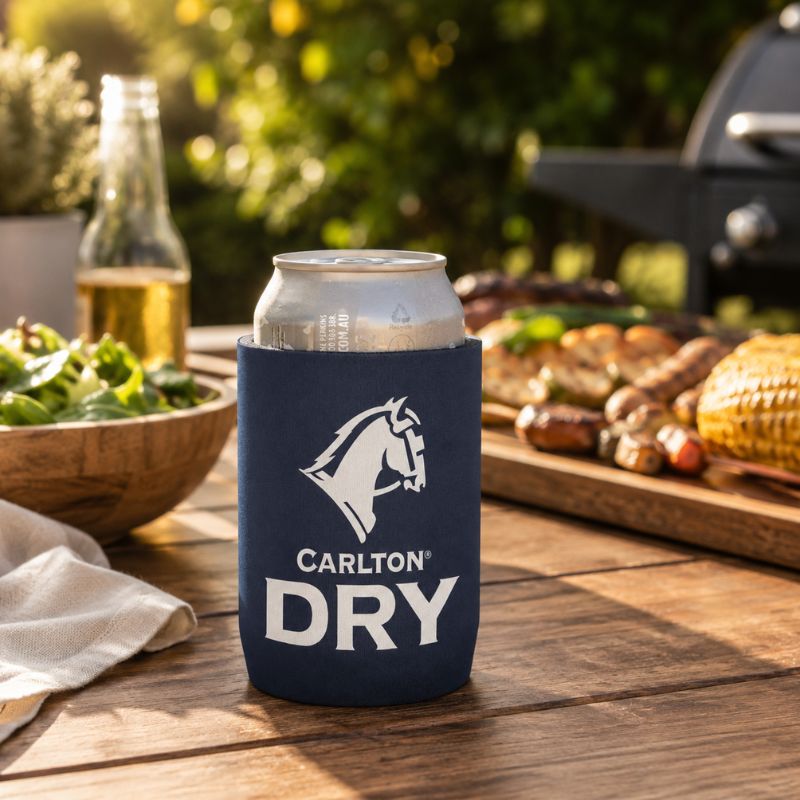 A navy blue Carlton Dry Classic Stubby Cooler with a white horse logo and the text 'CARLTON DRY' holding a silver can, set on a wooden outdoor table with a grill, salad, and corn in the background.