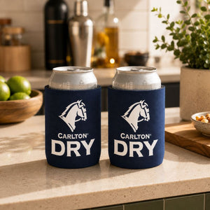 Two navy blue Carlton Dry Classic Stubby Cooler can coolers holding silver cans, set on a kitchen countertop with limes, bottles, and a plant in the background.