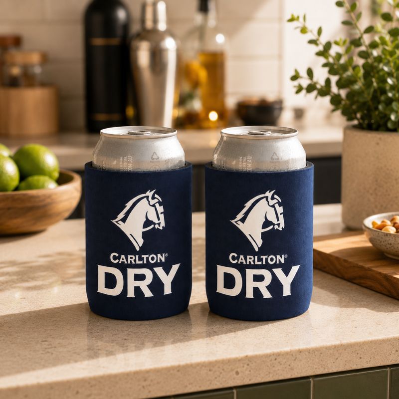 Two navy blue Carlton Dry Classic Stubby Cooler can coolers holding silver cans, set on a kitchen countertop with limes, bottles, and a plant in the background.