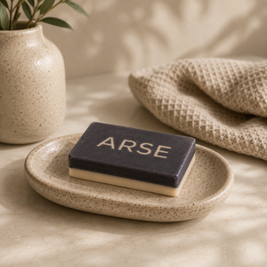 Bar of soap with 'ARSE' text on a speckled ceramic plate, next to a plant and towel.