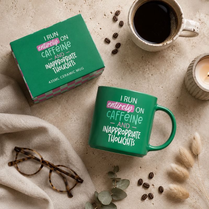 A Cheeky Caffeine Mug and its matching green box are arranged on a beige fabric surface with coffee, glasses, a candle, and dried grasses. The mug and box both read, 'I RUN entirely ON caffeine AND inappropriate thoughts' in white and pink lettering.