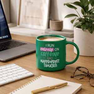 A Cheeky Caffeine Mug with white text reading 'I RUN entirely ON CAFFEINE AND INAPPROPRIATE THOUGHTS' sits on a wooden desk in a styled workspace setting with a laptop, keyboard, notebook, pen, and plant.
