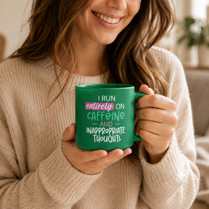 A woman in a beige knit sweater holds a Cheeky Caffeine Mug with the text 'I RUN entirely ON CAFFEINE AND INAPPROPRIATE THOUGHTS' in white, pink, and light green lettering.