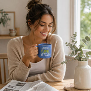 A woman in a cozy beige cardigan sits at a wooden table by a window, holding a blue Cheeky No Thanks Mug with a humorous quote printed in yellow and white text. A white vase with eucalyptus and an open newspaper are on the table beside her.