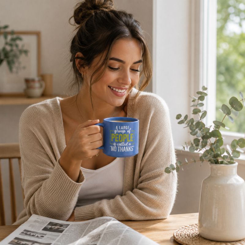A woman in a cozy beige cardigan sits at a wooden table by a window, holding a blue Cheeky No Thanks Mug with a humorous quote printed in yellow and white text. A white vase with eucalyptus and an open newspaper are on the table beside her.