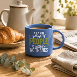 A blue ceramic Cheeky No Thanks Mug with a humorous quote printed on it, styled on a wooden table with a croissant, teapot, and newspaper in a warm, bright kitchen setting.