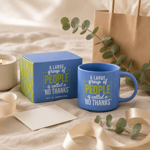 A blue Cheeky No Thanks Mug with a matching blue gift box sits on a beige fabric surface with ribbon, eucalyptus leaves, a candle, and a brown paper bag. The mug and box are printed with the phrase, 'A LARGE group of PEOPLE is called a "NO THANKS"'.