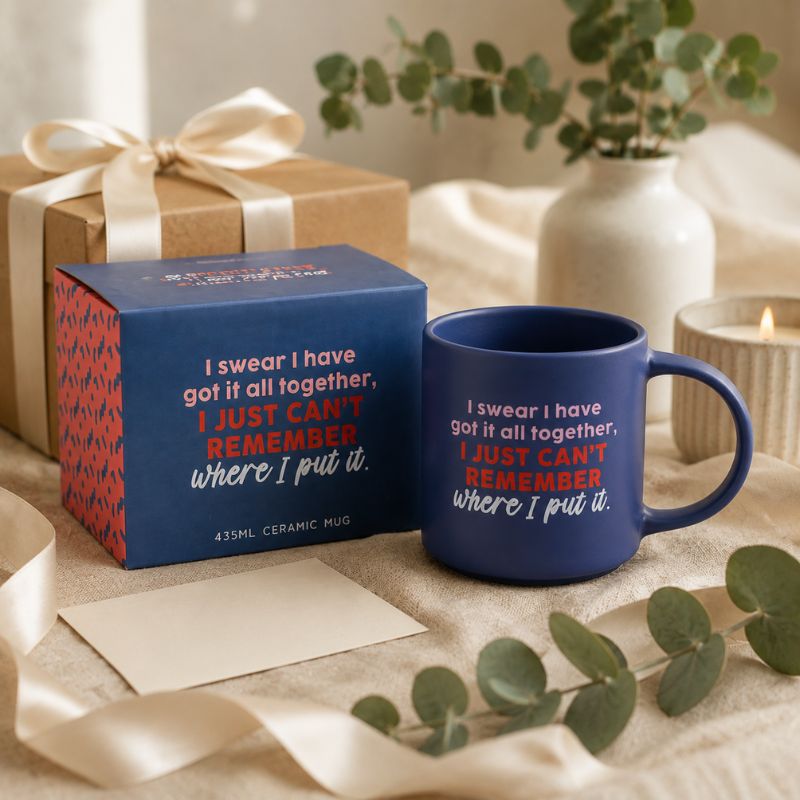A navy blue Cheeky Remember Mug with a humorous quote printed on it, displayed next to its blue and red patterned gift box. The scene is styled with a wrapped gift box, a lit candle, and eucalyptus leaves on a neutral fabric background.