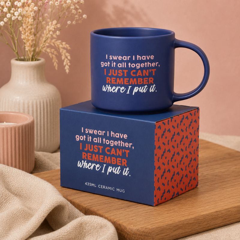 A Cheeky Remember Mug with a humorous quote sits on top of its matching blue and red patterned gift box, styled on a wooden board with a neutral background and dried flowers.