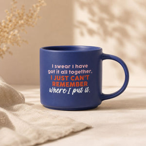 A dark blue cheeky remember mug with a humorous quote printed on it, styled on a beige fabric surface with soft shadows and dried flowers in the background.