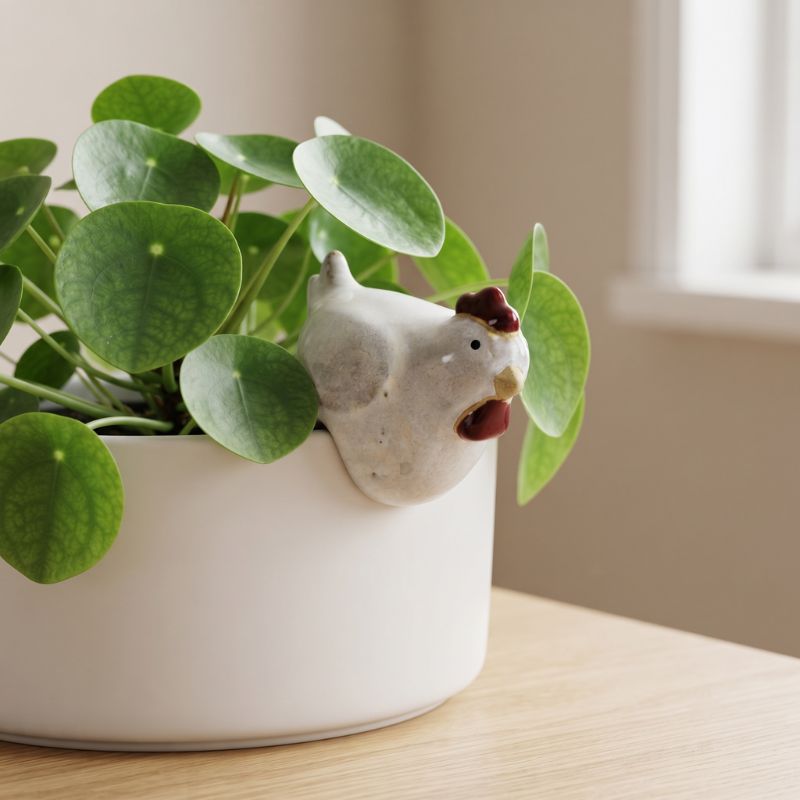 A white ceramic planter with a small white chicken figurine peeking out from behind round green plant leaves, placed on a light wood surface near a softly lit window.-Chicken Pot Hanger White 6cm