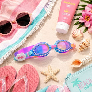 Cloud Blue Sunny Day Swim Goggles surrounded by sunscreen, tropical flowers, seashells, sunglasses, and beach essentials.
