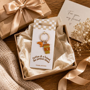 A Coffee and a Friend Keyring displayed on a branded card inside a gift box, surrounded by beige ribbon, tissue paper, and a card that reads 'For my friend'. The charm features a coffee cup and a small dog silhouette in warm orange, yellow, and brown tones.
