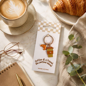 A coffee and a Friend Keyring displayed on a branded card, styled in a cozy flat-lay with a cup of latte, croissant, notebook, glasses, and greenery on a marble surface.