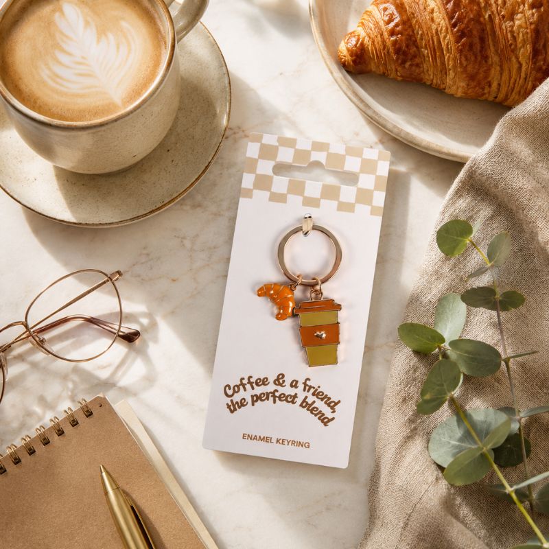 A coffee and a Friend Keyring displayed on a branded card, styled in a cozy flat-lay with a cup of latte, croissant, notebook, glasses, and greenery on a marble surface.