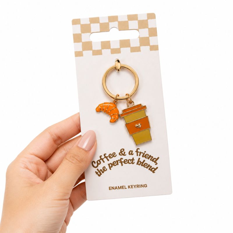 A hand holding a Coffee and a Friend Keyring on its retail display card. The keyring features a small orange croissant charm and a coffee cup charm with a gold-tone metal ring.