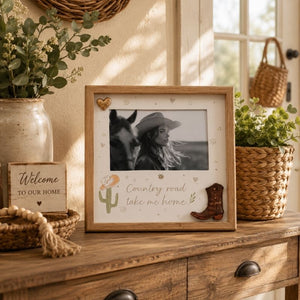Decorative framed Country Road Take Me home picture with a cowboy theme on a wooden surface with plants and a 'Welcome to our home' sign.