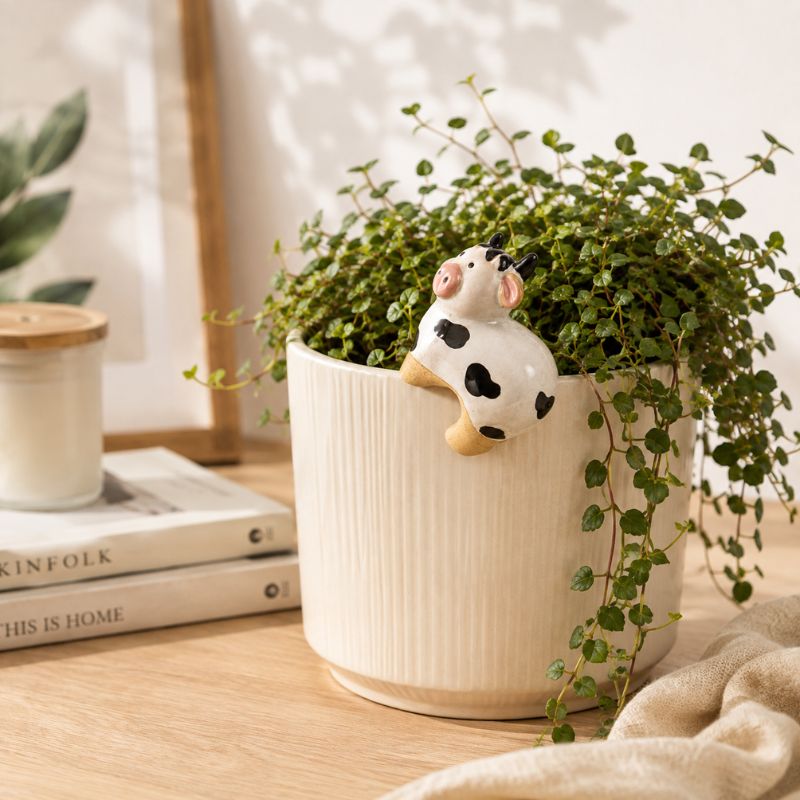 A cream-colored ribbed ceramic planter holding a trailing green plant, with a small black-and-white cow figurine perched on the rim, styled on a wooden tabletop with books and a candle in the background.-Cow Pot Hanger - 8cm