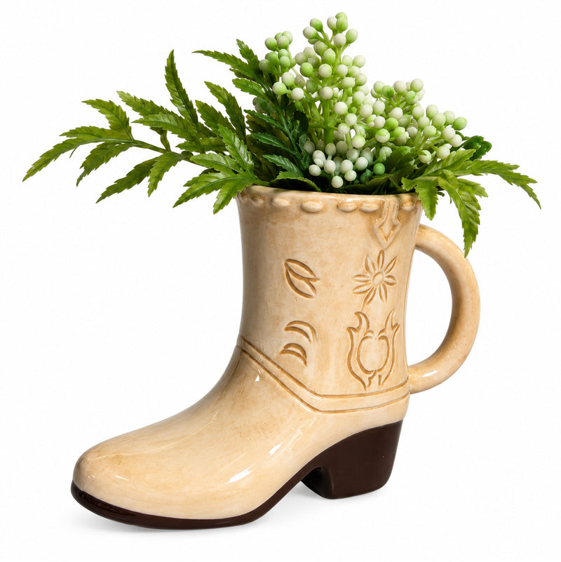Tan ceramic cowboy boot mug styled with green foliage on a white background.