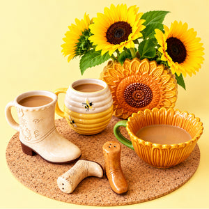 Tan ceramic cowboy boot mug styled with sunflower-themed drinkware, bright sunflowers and Western accessories on a cork mat.