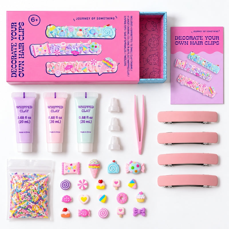 A children's craft kit laid out on a white background, including a pink box labeled 'Journey of Something' with hair clips, three small tubes of whipped clay, tweezers, pink hair clip bars, a bag of multicolor sprinkles, and assorted pastel decorative charms.