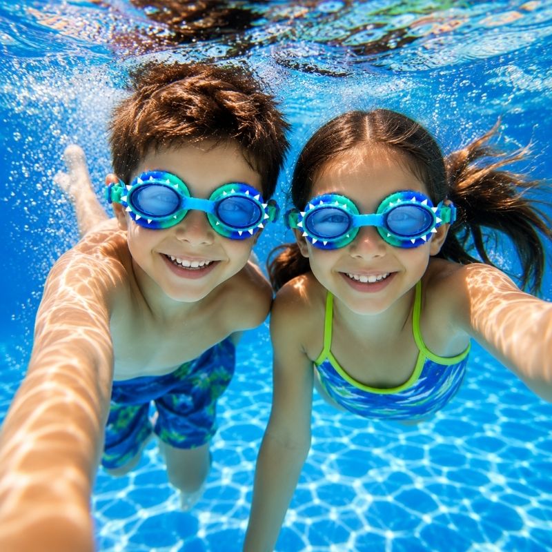 Boy and girl swimming underwater wearing Dino Royal dinosaur swim goggles in a bright blue pool